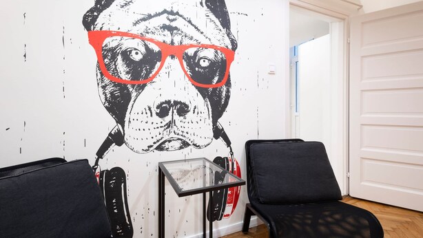 A room with a wall mural of a dog wearing glasses, two black chairs, a small table, and a white door.