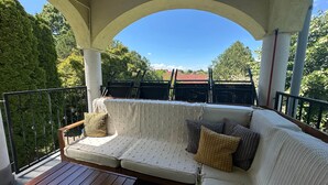 A covered outdoor seating area with a cushioned bench, a woven basket, and a view of the surrounding greenery and buildings. - LB - Art Gallery Apartman (Siófok)