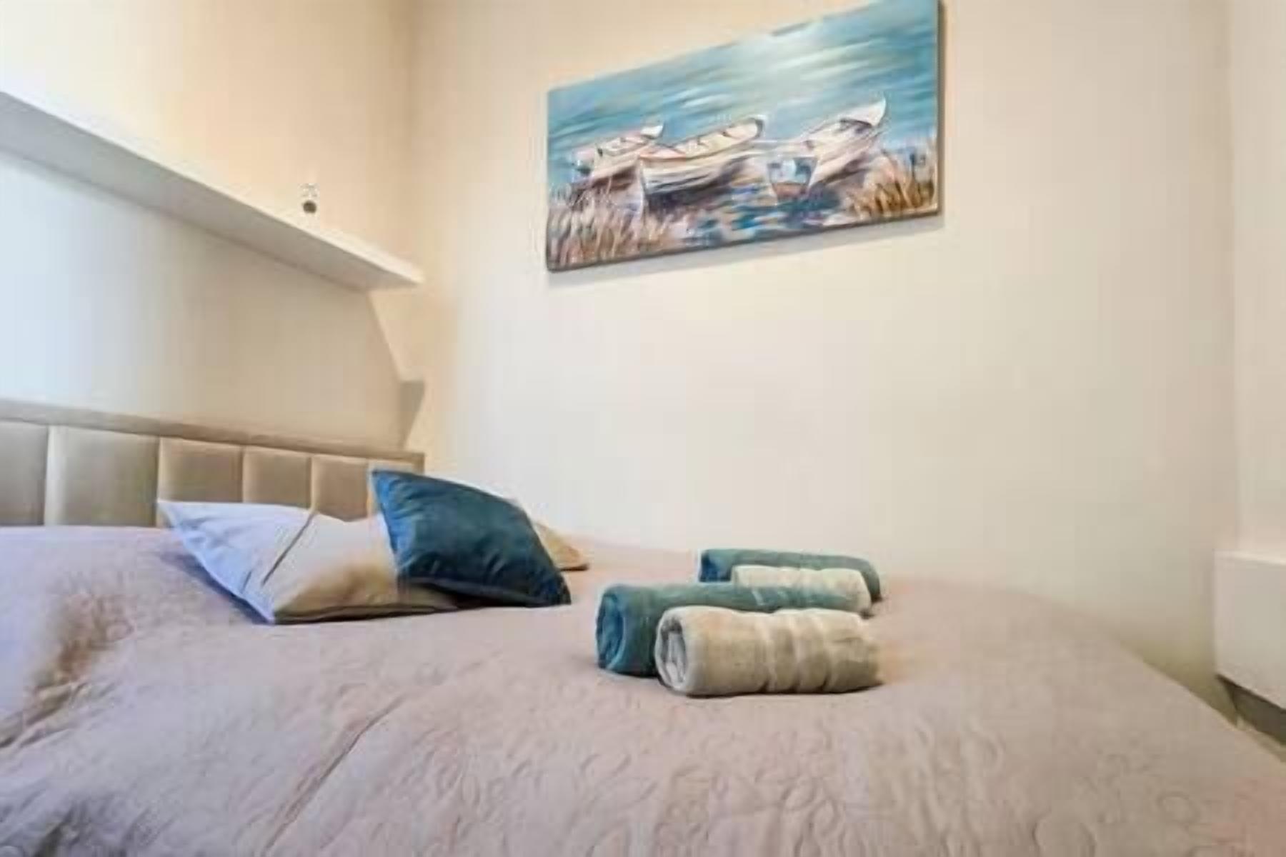 A neatly made bed with a headboard, a painting of boats on water, and rolled towels on the bed.