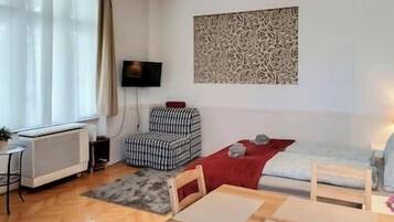 A compact living space with a sofa, a chair, a small table, a radiator, a TV, and a patterned wall decoration.