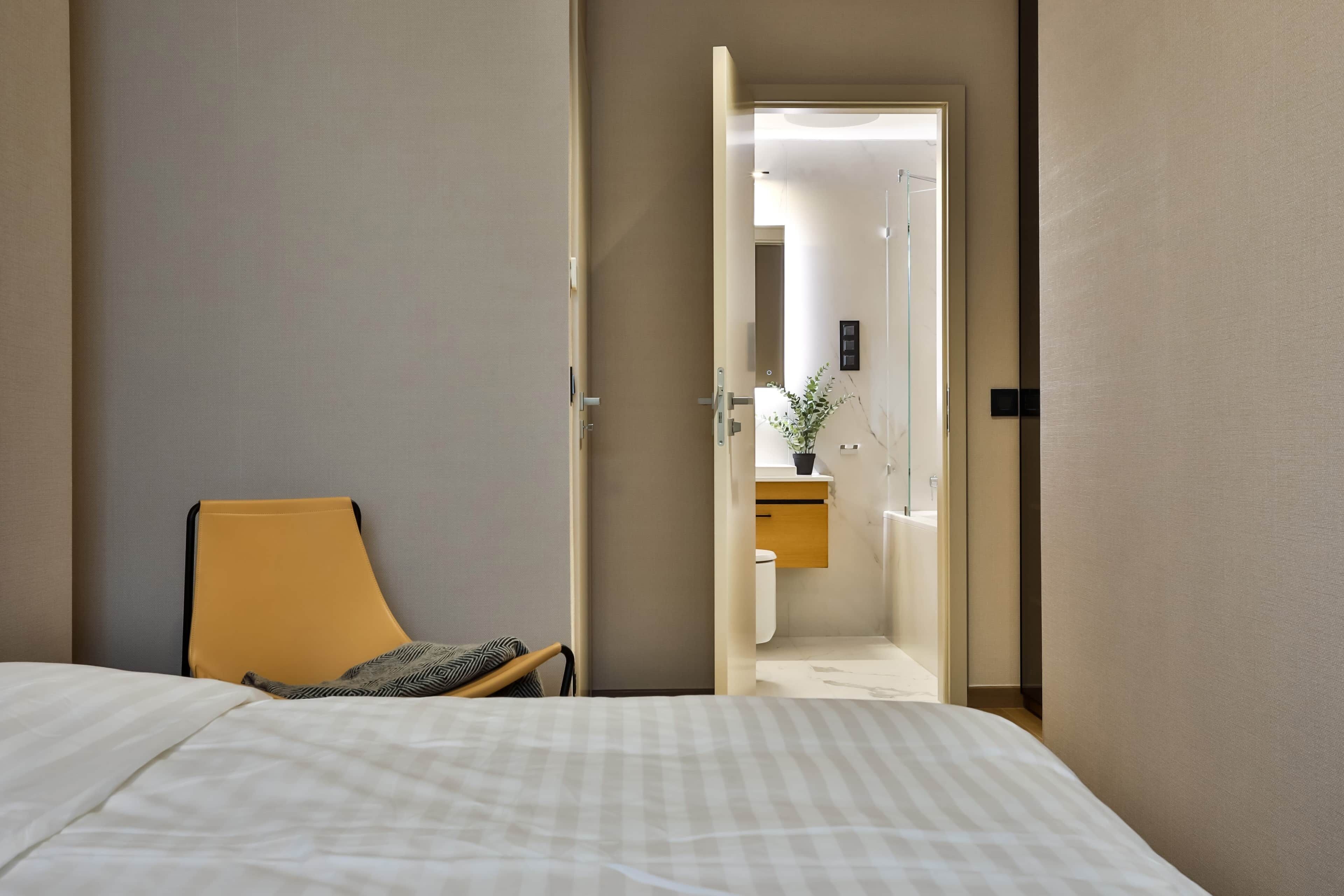 Ein Hotelzimmer mit Bett, Stuhl, einem durchgegangenen Tür erkennbaren Badezimmer und einem Blumentopf auf der Badezimmerablage.
