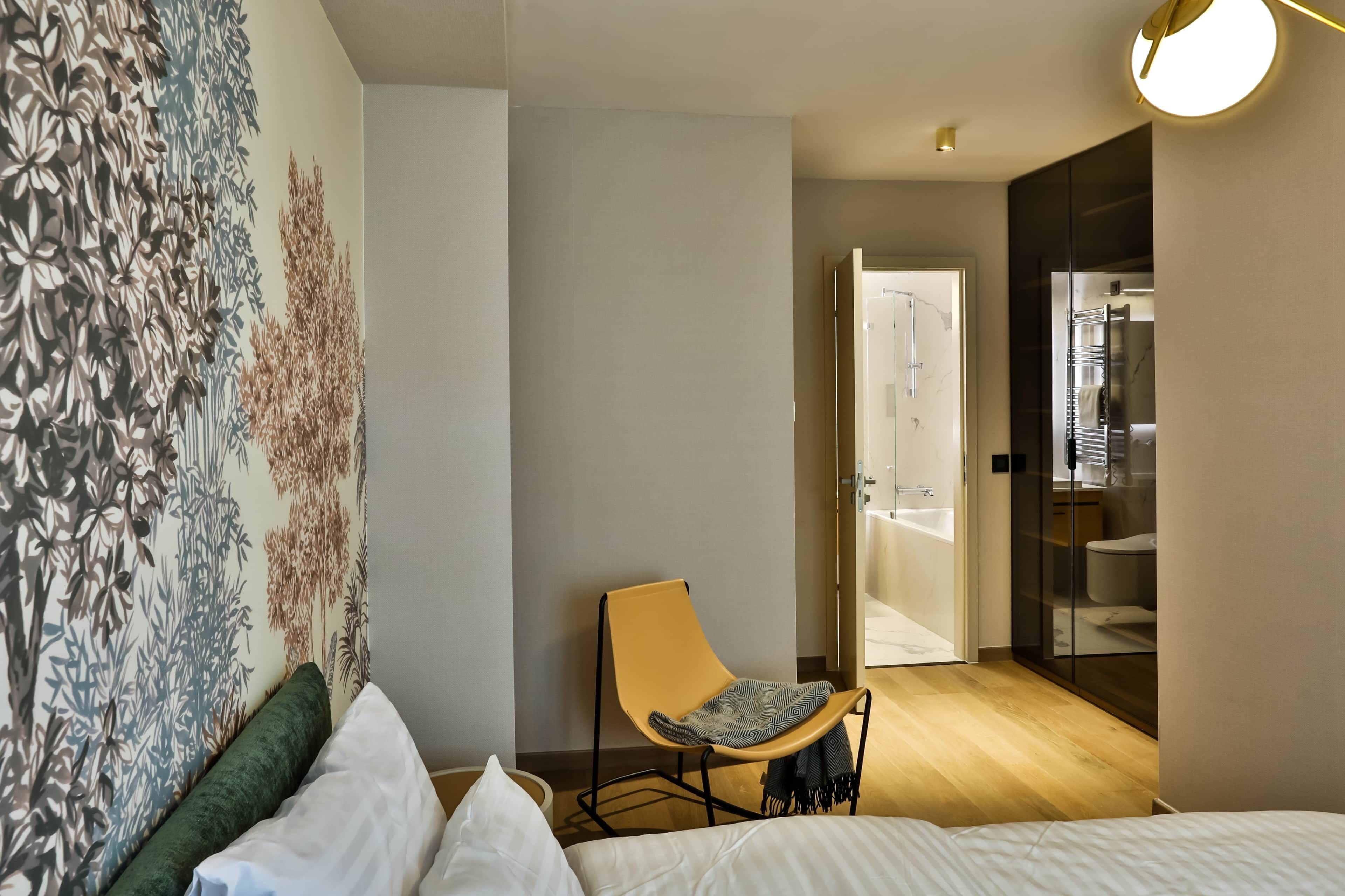 Habitació d'hotel moderna amb un llit, una cadira, un mural gran a la paret i un bany visible a través d'una porta oberta.