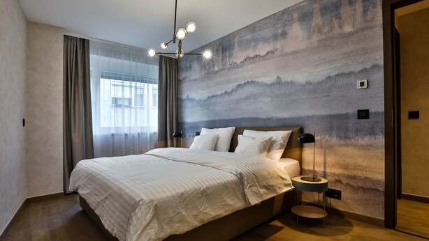 A modern bedroom with a large bed, a wall-mounted light fixture, and a view of the outdoors through a window.