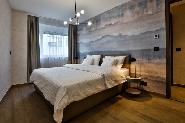 A modern bedroom with a large bed, a wall-mounted light fixture, and a view of the outdoors through a window.