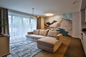 A modern living room with a sofa, a dining table with chairs, a large window with sheer curtains, and a wall mural.