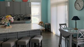 A modern kitchen with a central island, bar stools, a dining table with chairs, and a clock on the wall.