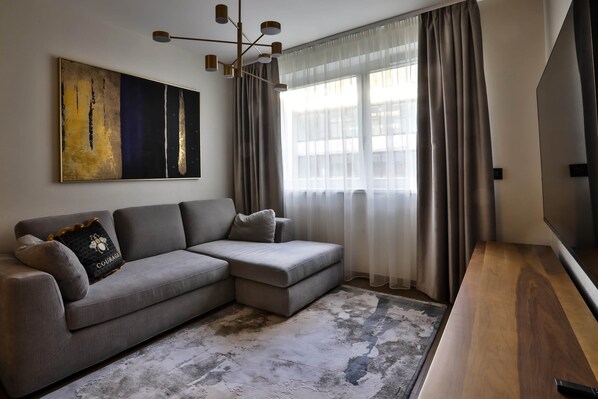 A modern living room with a grey sectional sofa, a large abstract painting, a contemporary chandelier, and a large window with sheer curtains.