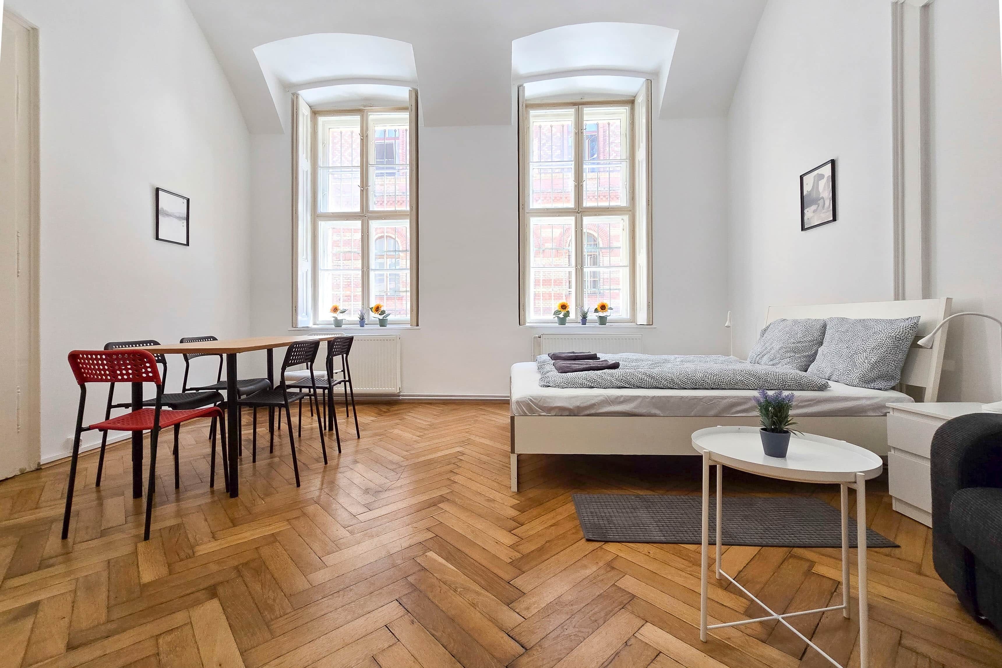 A modern apartment with a herringbone wooden floor, a white sofa, a small round table, and two windows with a view of a brick building.