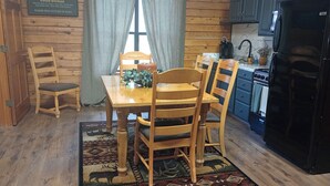 Een houten hut interieur met een eettafel, stoelen, een keukenhoek en een bord waarop staat 'WANTED GOOD WOMAN'.