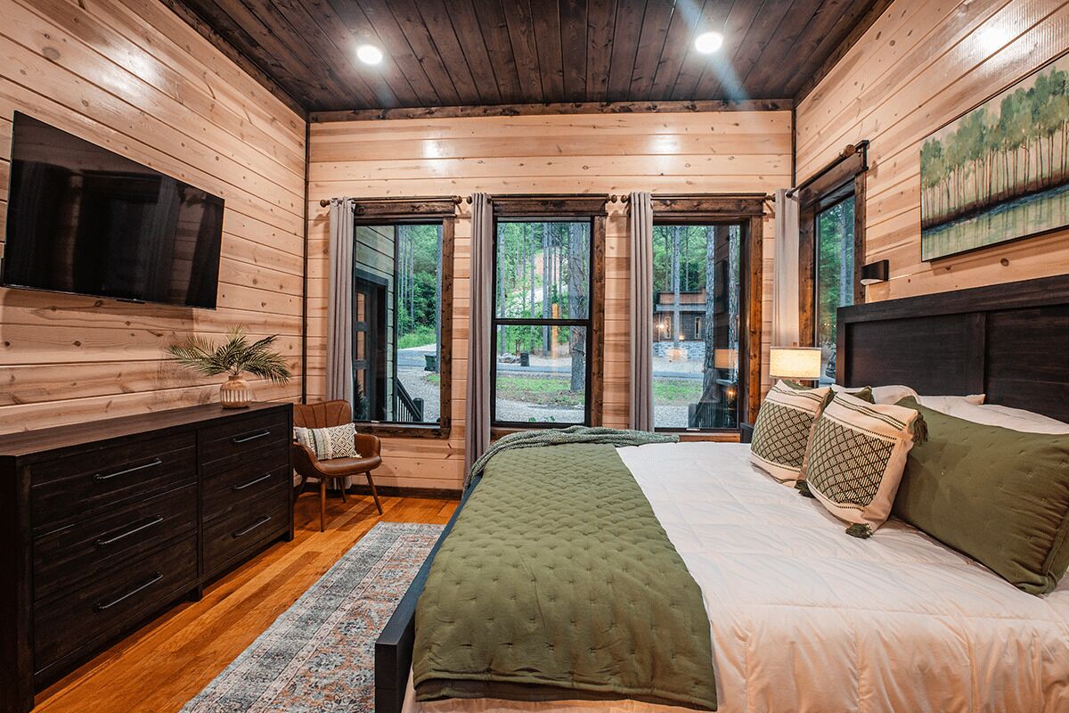 Ein Schlafzimmer einer Holzhütte mit Bett, Schminktisch, Fernseher und großen Fenstern.