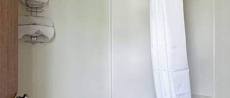 Een badkamer met een toilet, een douche met een witte gordijn en een stap opgevouwen handdoeken aan de muur.