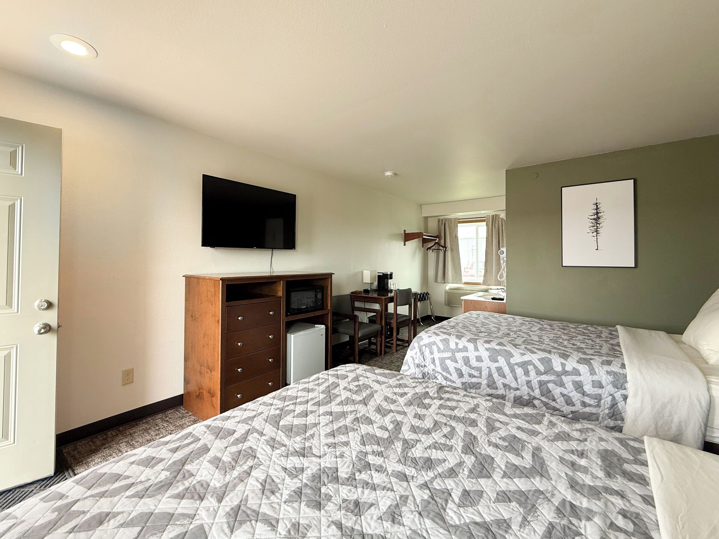 Ein Hotelzimmer mit zwei Betten, einem Schreibtisch mit Stuhl, einem Fernseher, einer Mikrowelle, einem kleinen Kühlschrank, einem Badezimmer mit Waschbecken und Spiegel sowie einem gerahmten Bild an der Wand.