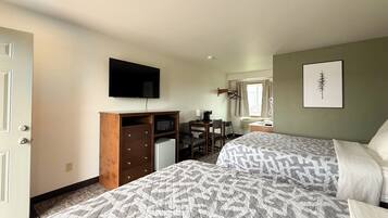 Ein Hotelzimmer mit zwei Betten, einem Schreibtisch mit Stuhl, einem Fernseher, einer Mikrowelle, einem kleinen Kühlschrank, einem Badezimmer mit Waschbecken und Spiegel sowie einem gerahmten Bild an der Wand.