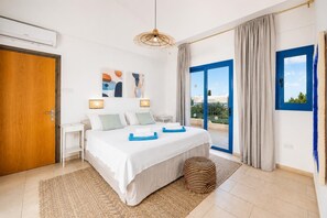 A bedroom with a bed, bedside tables, a hanging light fixture, a view through a window, and a door.