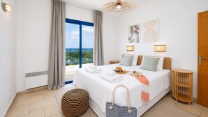 A hotel room with a large bed, a view of the sea, a round ottoman, and a wooden side table.