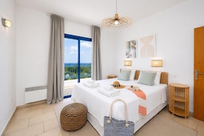 Camera d'hotel con un letto grande, vista sul mare, un poggiapiedi rotondo e un tavolino in legno.