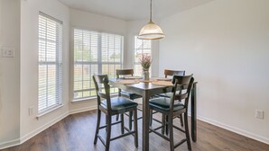 Comedor con mesa de madera, cuatro sillas, lámpara colgante y ventanas grandes con persianas.