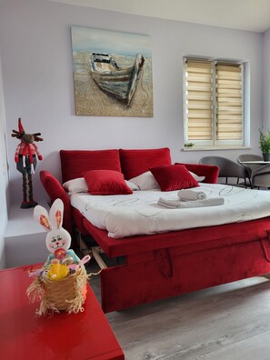 Een slaapkamer met een rood bed, een schilderij van een boot aan de muur en een raam met jaloezieën.