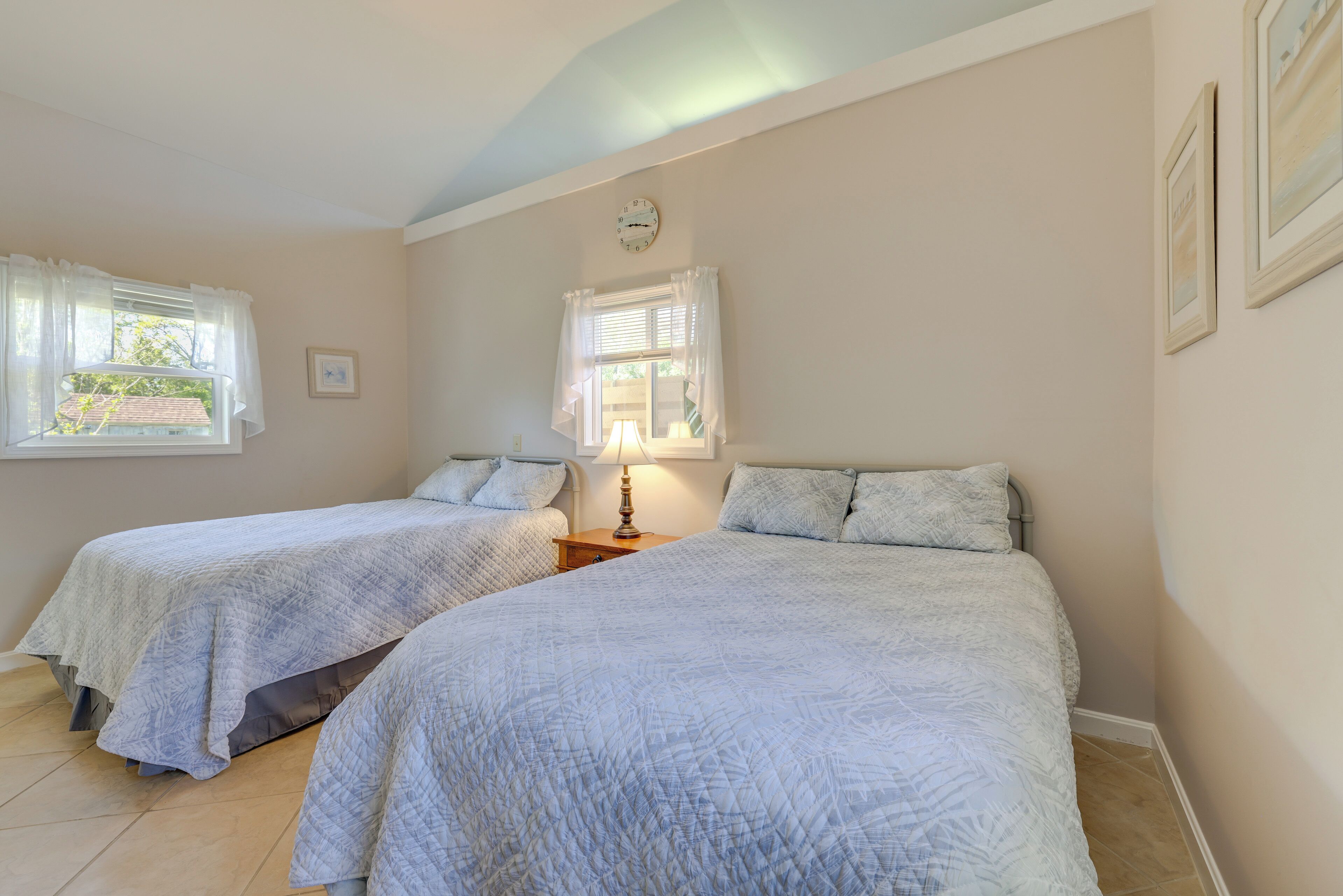 A bedroom with two beds, each with a white quilted bedspread, a bedside table with a lamp, and a window with white curtains.