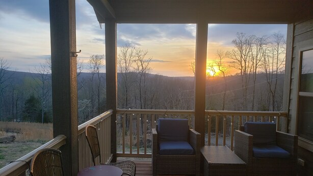 A wooden deck with wicker furniture and a table, overlooking a forested landscape with a sunset in the background.