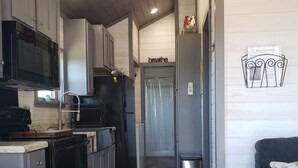 A narrow kitchen with wooden flooring, a black refrigerator, a microwave, and a sink. A grey sofa is visible in the background.