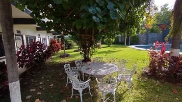 A garden area with a table and chairs, surrounded by greenery and a pool in the background.