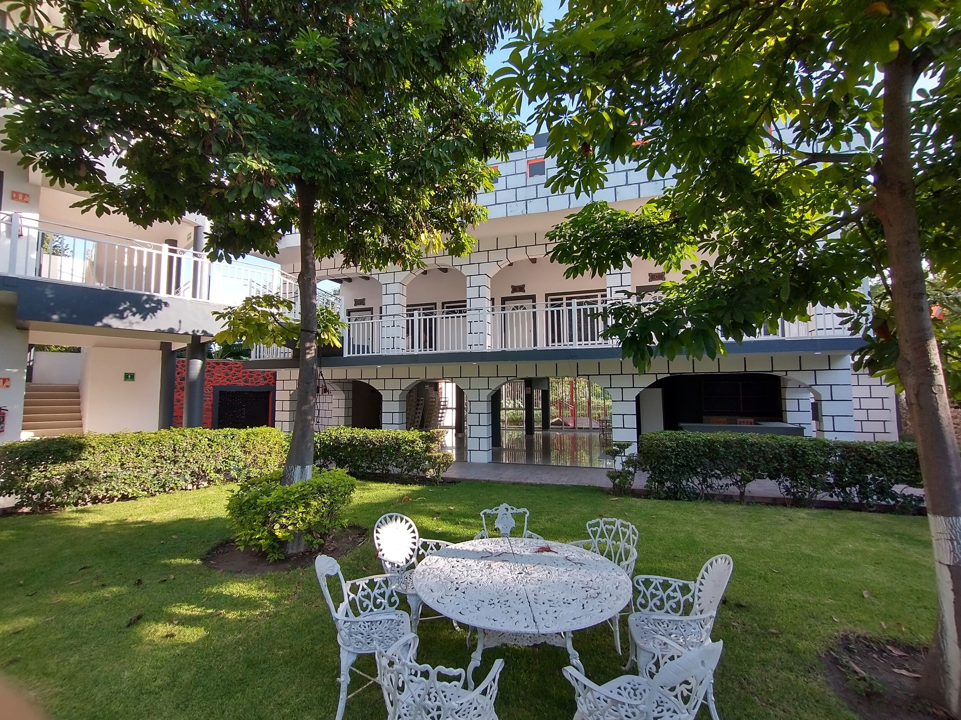 Un patio con una mesa redonda y sillas, rodeado de vegetación y un edificio con balcón.