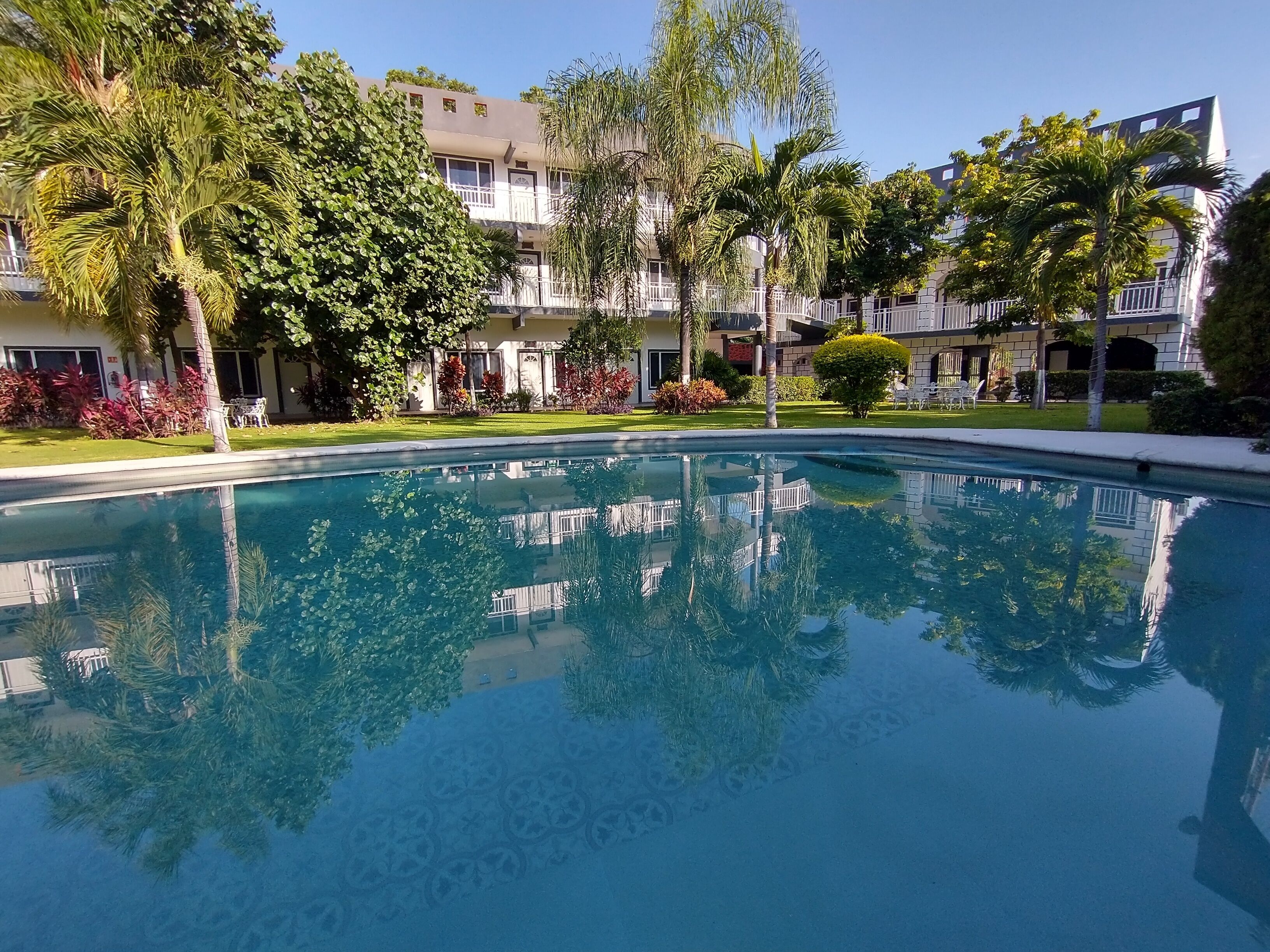 A clear swimming pool surrounded by lush greenery and multi-story buildings with balconies.