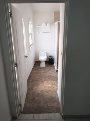 Un pasillo estrecho que conduce a un baño con inodoro, un cubo de basura y un estante con toallas.