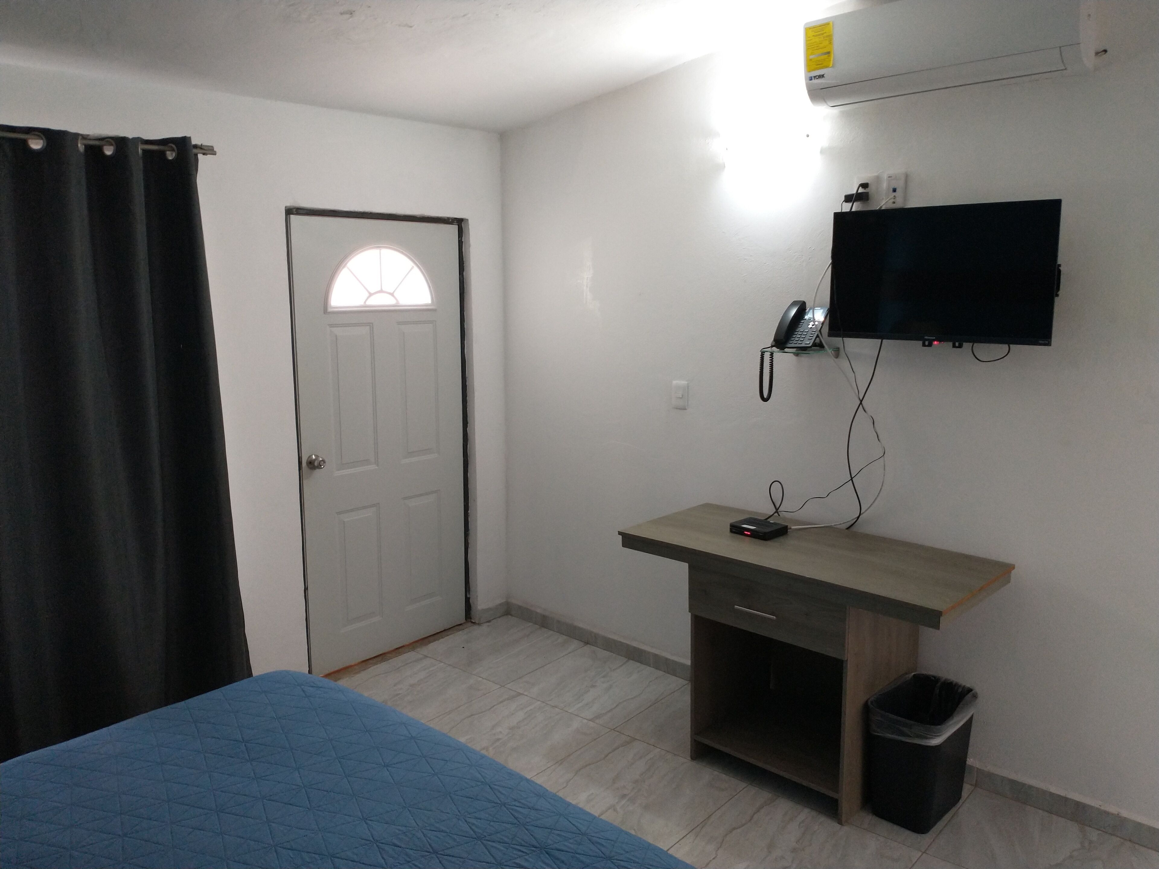 A room with a bed, a desk with a computer, a trash bin, a wall-mounted phone, and a TV.