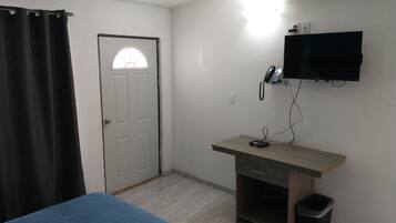 A room with a bed, a desk with a computer, a trash bin, a wall-mounted phone, and a TV.