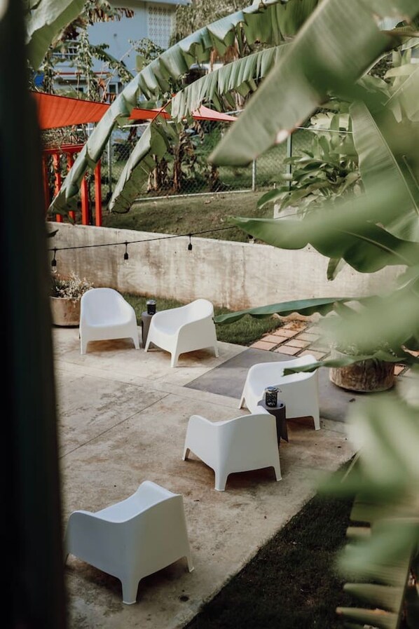 An outdoor seating area with white plastic chairs and a concrete floor, surrounded by greenery and a red fence.