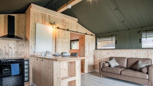 A compact kitchen with a stove, sink, and wooden cabinetry. A sofa and a window are visible in the living area. - Paviland - Safari Glamping Tent - Llangennith (Llangennith)