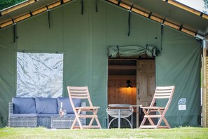 Een gezellig buitenverblijf met een groene tentconstructie, een rieten bank, twee houten stoelen en een kleine ronde tafel.