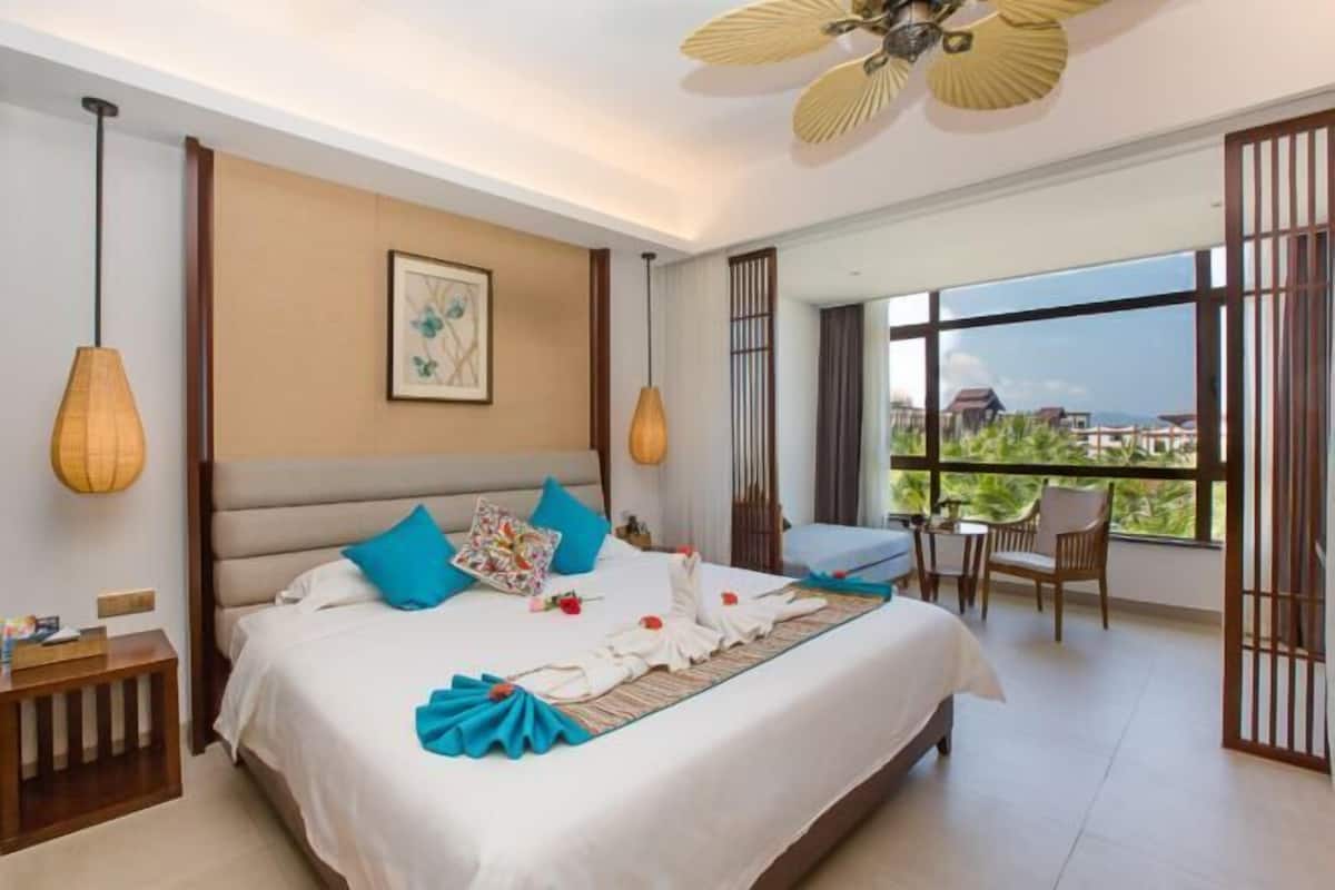 Quarto de hotel com uma cama grande, um ventilador de teto, vista para o exterior e uma pequena mesa com cadeiras.