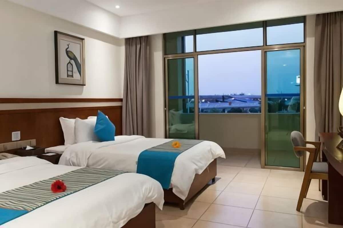 Quarto de hotel com duas camas, uma mesa, uma cadeira e uma janela grande com vista.