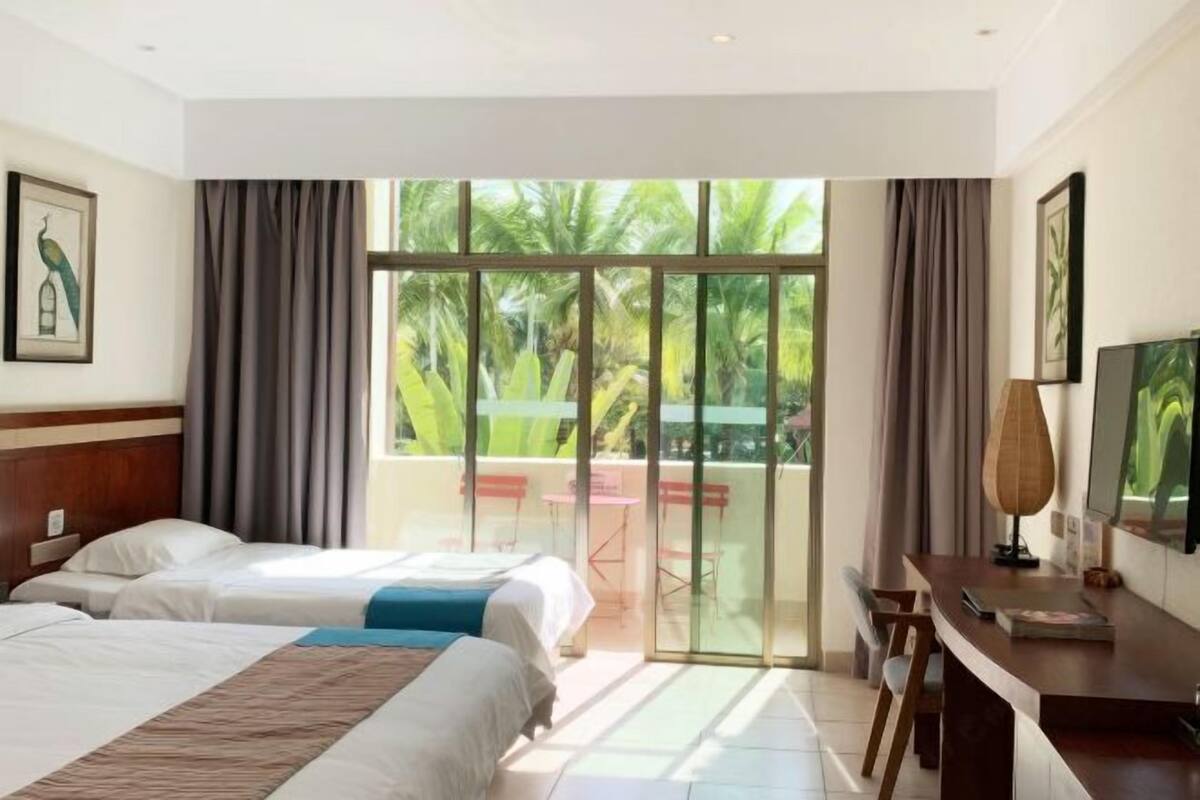 Quarto de hotel com duas camas, uma escrivaninha, uma cadeira e uma porta de vidro deslizante para área externa com mesa e cadeiras.
