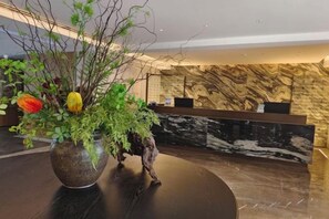 A reception desk with a marble wall backdrop, a wooden reception counter, and a large vase with green foliage and colorful flowers.