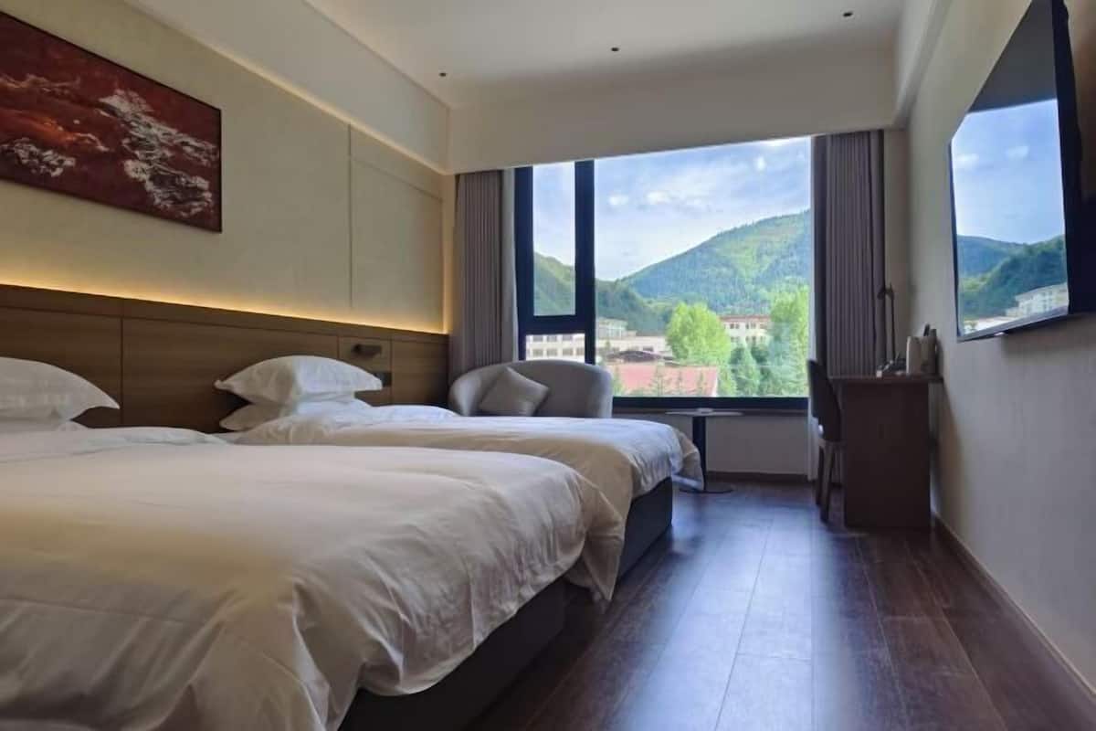 Quarto de hotel com duas camas, uma escrivaninha, uma cadeira e uma janela grande com vista para as montanhas.