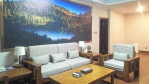A modern hotel room with a large painting of a forest and lake, a grey sofa, two armchairs, a wooden coffee table, and a small side table with a lamp.
