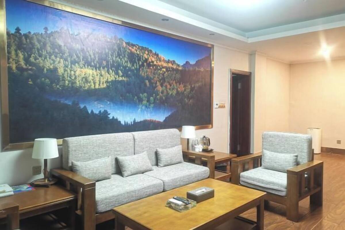 Quarto de hotel moderno com uma grande pintura de uma floresta e um lago, um sofá cinzento, duas poltronas, uma mesa de centro de madeira e uma pequena mesa lateral com uma luminária.