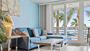 Un salon avec un canapé bleu, une table basse en bois et une lampe. Une grande fenêtre offre une vue sur des palmiers, et il y a un balcon.