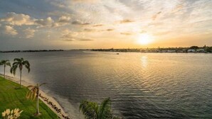 Un coucher de soleil sur une étendue d’eau calme, bordée d’une rive herbeuse et de palmiers.