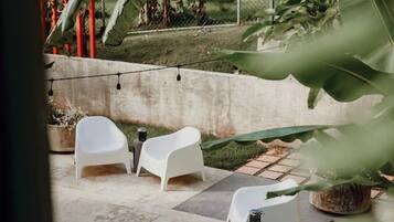 An outdoor seating area with white plastic chairs and a concrete floor, surrounded by greenery and a red fence.