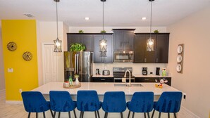 A modern kitchen with dark cabinets, a central island with blue bar stools, stainless steel appliances, and pendant lighting. - Go 28 Champions Gate (Davenport)