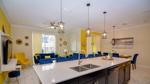 A modern kitchen with a central island, black cabinets, and a white countertop. There are blue chairs and a yellow sofa in the adjacent living area. - Go 28 Champions Gate (Davenport)