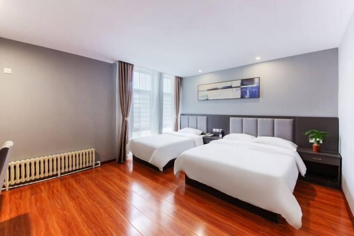 Quarto de hotel com duas camas, um radiador, uma janela grande com cortinas, uma obra de arte na parede e uma pequena planta.