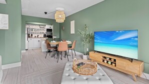 A modern living room with a large flat-screen TV, a dining area with a table and chairs, and a kitchen in the background. - Private Garden Apartment with Heated Pool 1BR (Sarasota)