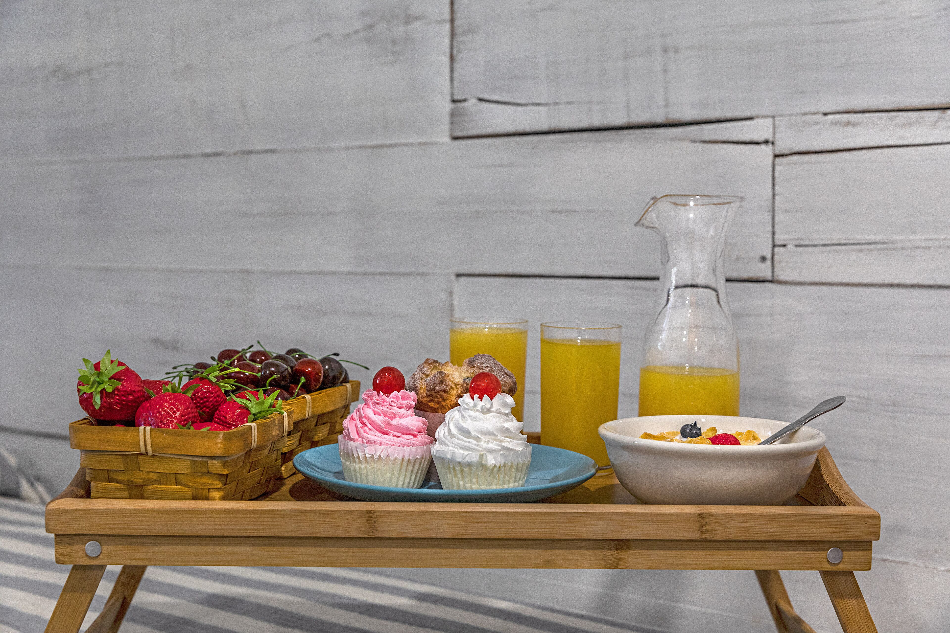 A wooden tray with a basket of strawberries, two cupcakes, a bowl of cereal, and two glasses of orange juice.