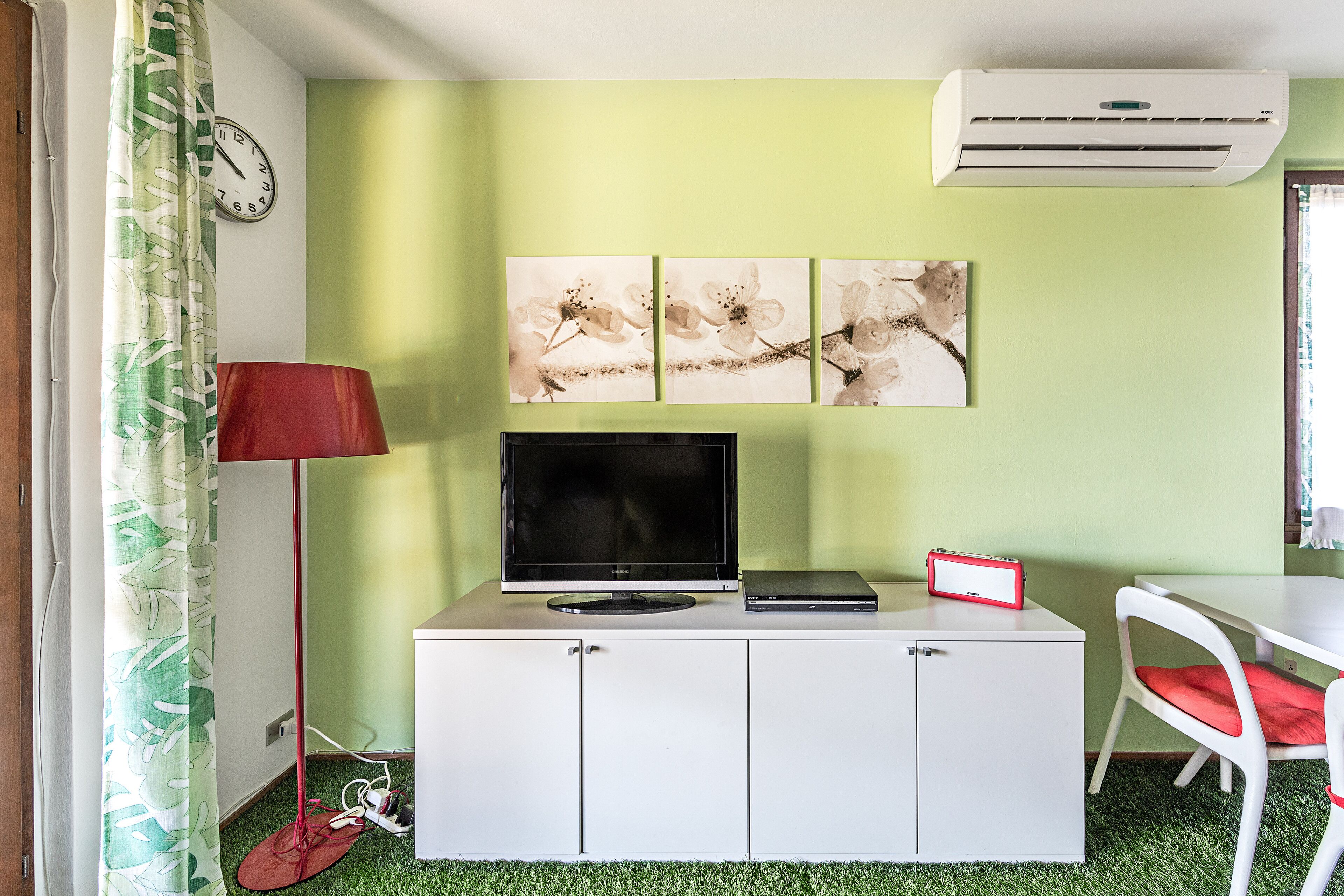 Ein Zimmer mit einem weißen Schrank, einem Flachbildfernseher, einer roten Lampe und einem roten Sessel. An der Wand hängen drei Gemälde und über dem Schrank befindet sich eine Klimaanlage.